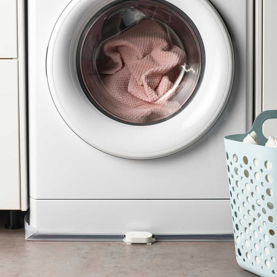 Weißer IKEA KLIPPBOK Wasserlecksensor auf dem Boden vor einer Waschmaschine neben einem Wäschekorb